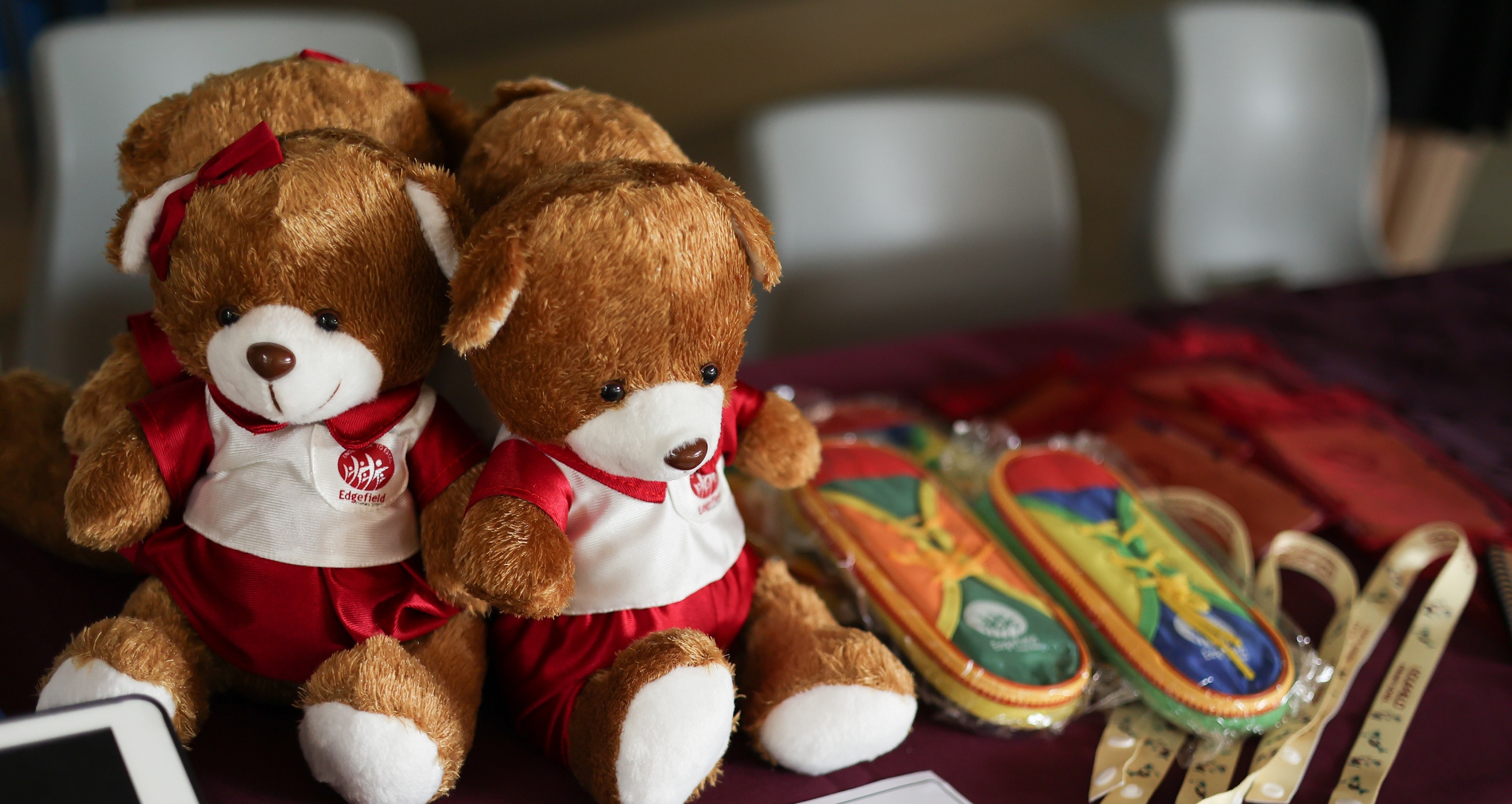 Two brown teddy bears in red and white outfits sit on a table beside colorful slippers and ribbon.